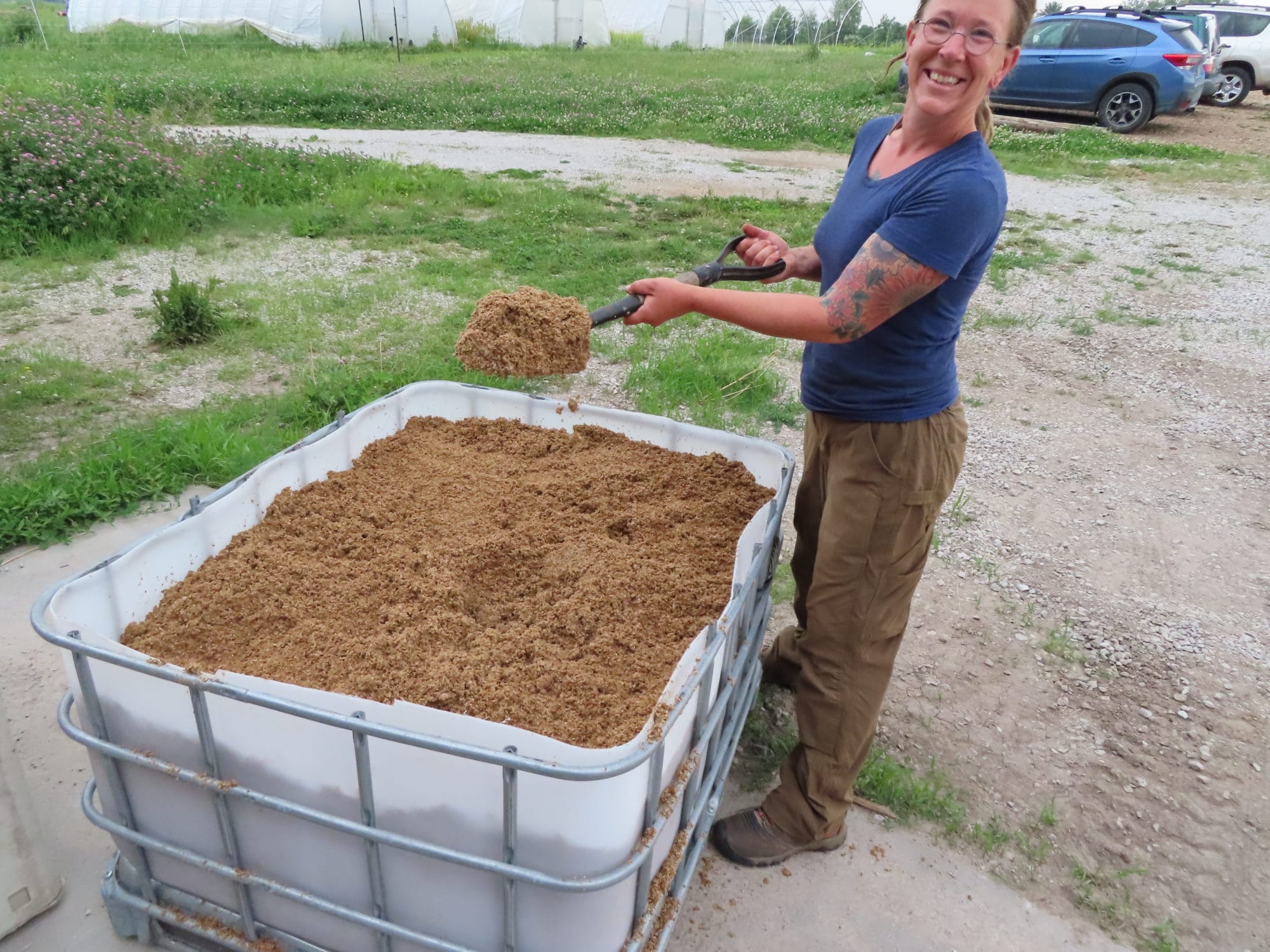 Kate Solko standing next to a big bin of brewers spent grain. She holds a shovel full of grain in her hands.